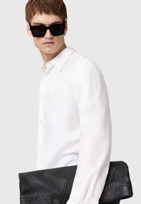 Camisa blanca de botones con cuello clásico, clutch de cuero negro texturizado y gafas de sol grandes negras. La modelo posando contra un fondo gris.