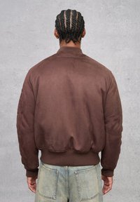 Giacca bomber marrone con una texture liscia, polsini e orlo a costine, e un piccolo logo ricamato sulla schiena, indossata sopra un jeans denim blu chiaro.