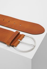 Ceinture en cuir brun avec une boucle en métal argenté ronde, dotée de trous pour ajustement et d'une finition lisse et texturée.