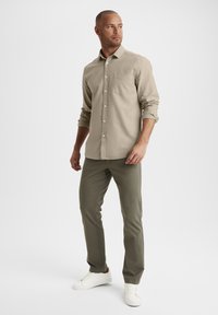 Beige Hemd mit Knöpfen und einer Brusttasche, kombiniert mit olivgrünen Hosen und weißen Sneakers. Glattes Material und taillierte Passform.