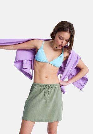 Jasnoniebieski trójkątny top bikini, zestawiony z luźną zieloną spódnicą na sznurku. Na jednym ramieniu spoczywa jasnofioletowa kurtka z fakturowanej tkaniny.