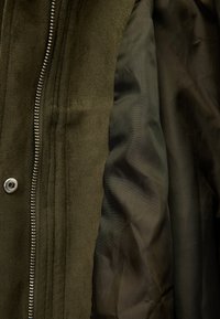 Chaqueta de ante verde oliva con un forro interior suave, que presenta un cierre de cremallera plateada y un botón a presión. Superficie exterior texturizada.