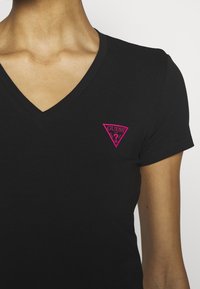 Czarny t-shirt z dekoltem w serek, wykonany z bawełny, z krótkimi rękawami, z różowym trójkątnym logo "GUESS" na lewej stronie klatki piersiowej.