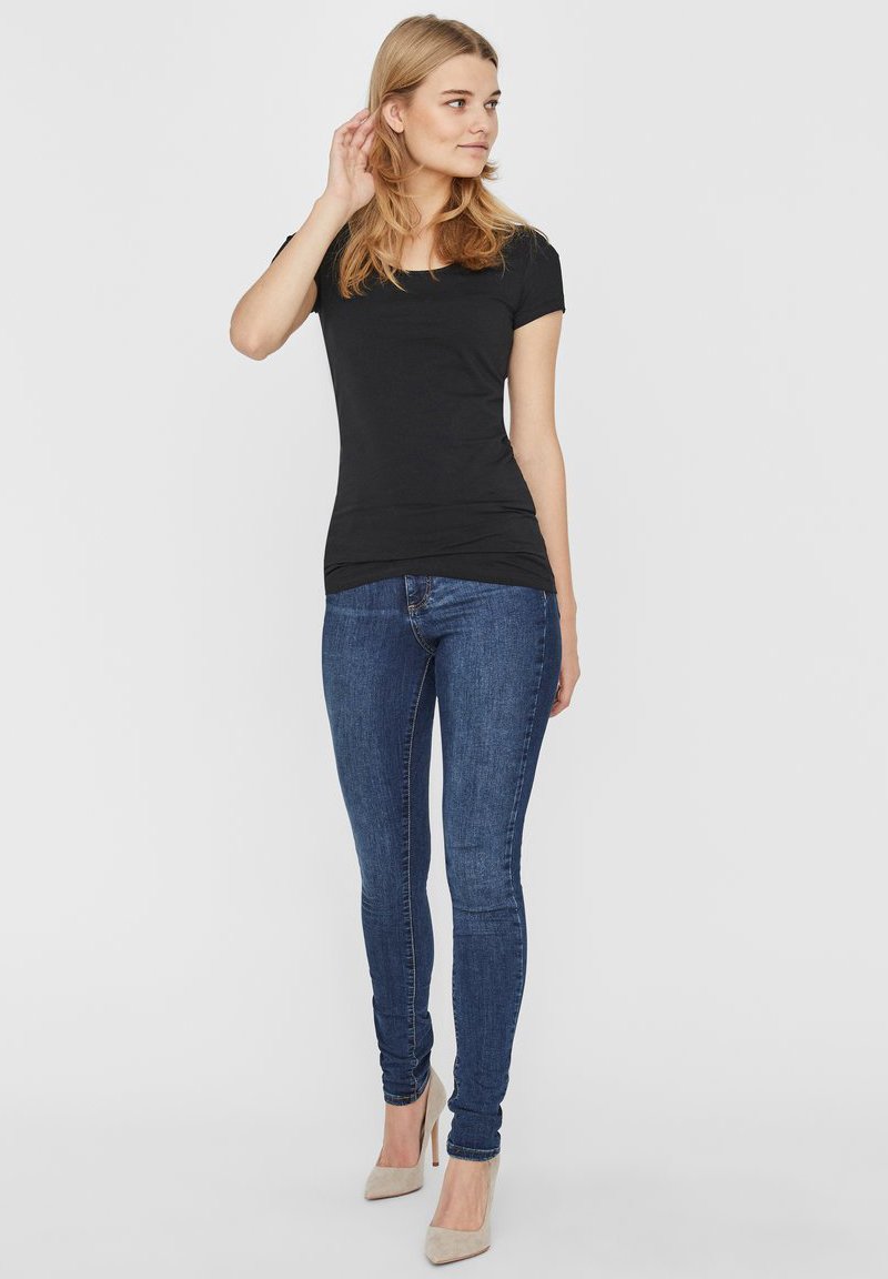 Sort tætsiddende t-shirt med rund hals, parret med mørkeblå skinny jeans. Model iført nude højhælede sko. Ensfarvet baggrund.