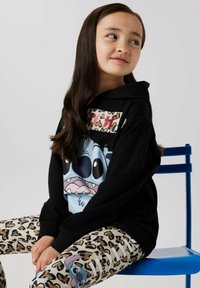 Svart huvtröja med stor blå tecknad karaktärsgrafik och djurmönstrade accenter, matchad med beige leggings som har ett brunt leopardmönster.