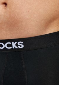 Black boxer shorts with a textured waistband featuring the word "STOCKS" in white. Smooth fabric with flat seams, focused on the waistband area.