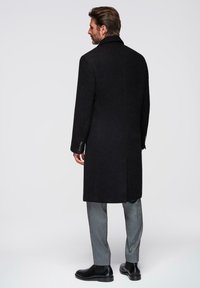 Abrigo de lana negro con cuello de muesca y abertura trasera, con puños abotonados, combinado con pantalones grises y botas de cuero negras.