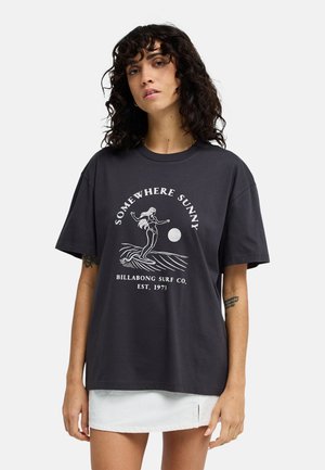 Frau mit lockigem dunklem Haar, die ein lockeres schwarzes T-Shirt mit Surf-Grafik und weißem Text trägt, kombiniert mit einem kurzen weißen Rock.