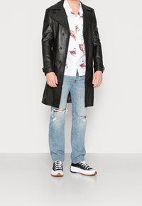 Czarny skórzany płaszcz trencz z podwójnym zapięciem na guziki, na kolorowej, wzorzystej koszuli. Jasnoniebieskie, postrzępione dżinsy i czarne trampki.