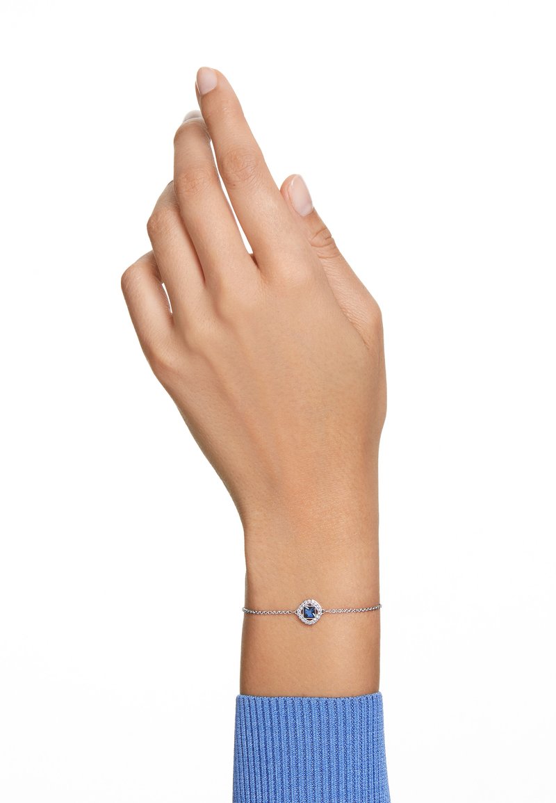 Srebrna zapestnica s osrednjim modrim kamnom, okrogle oblike in nežno verigo, nosijo na roki blizu teksturirane modre pulover.