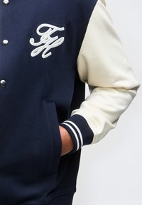 Άτομο που φοράει μπλε ναυτικό και κρεμ varsity μπουφάν με μονόγραμμα FH κεντημένο στο στήθος, με το χέρι στην τσέπη.