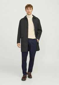 Μαύρο παλτό με κουμπιά, γιακά και πλαϊνές τσέπες, φορεμένο πάνω από μπεζ ζιβάγκο, συνδυασμένο με σκούρο μπλε παντελόνι και καφέ παπούτσια.