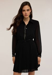 Schwarzes Faltenkleid mit langen durchsichtigen Ärmeln, Knopfleiste, zwei Brusttaschen und einem taillierten Bund mit dekorativem Besatz.