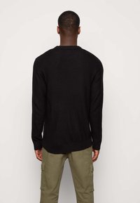 Homme portant un pull noir en maille et un pantalon cargo vert olive, debout face à un mur uni de couleur claire.