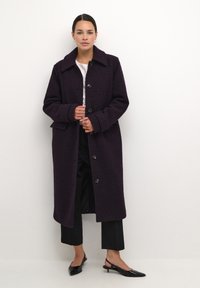 Cappotto in lana viola, con texture, grande colletto e chiusura frontale con bottoni. Indossato sopra un top bianco e pantaloni neri, con scarpe a punta.