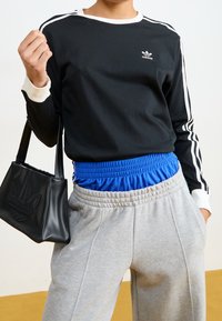 Černý top s dlouhým rukávem s bílými doplňky a logem, šedé tepláky s modrým elastickým pasem a černá taška s logem.