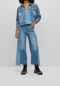 Modrá denimová bunda přes odpovídající modré tričko, v kombinaci s širokými modrými džíny s rozparkem, doplněná černými šněrovacími botami.