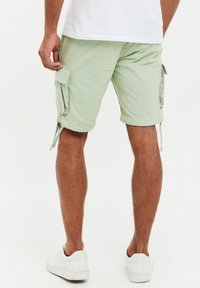 Hombre con pantalones cortos cargo de color verde claro y zapatillas blancas de pie contra un fondo blanco, vista desde atrás.