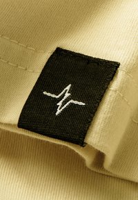 Cremfarbener Stoff mit glatter Textur; schwarzes Kleidungsetikett mit aufgesticktem weißen Blitzlogo an der Seite.