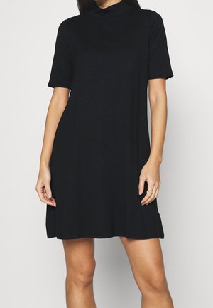 Femme portant une robe noire côtelée à manches courtes avec un col montant, debout devant un fond clair uni.