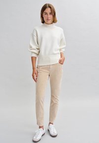 Lässiger weißer Strickpullover mit hohem Kragen, kombiniert mit hellbeigen Cordhosen und weißen Sneakers mit braunen Sohlen.
