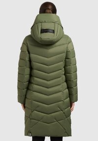 Olivengrøn vatteret jakke med chevronmønster, hætte og et sort mærke på ryggen. Har lommer med lynlås og en mellemlang længde.