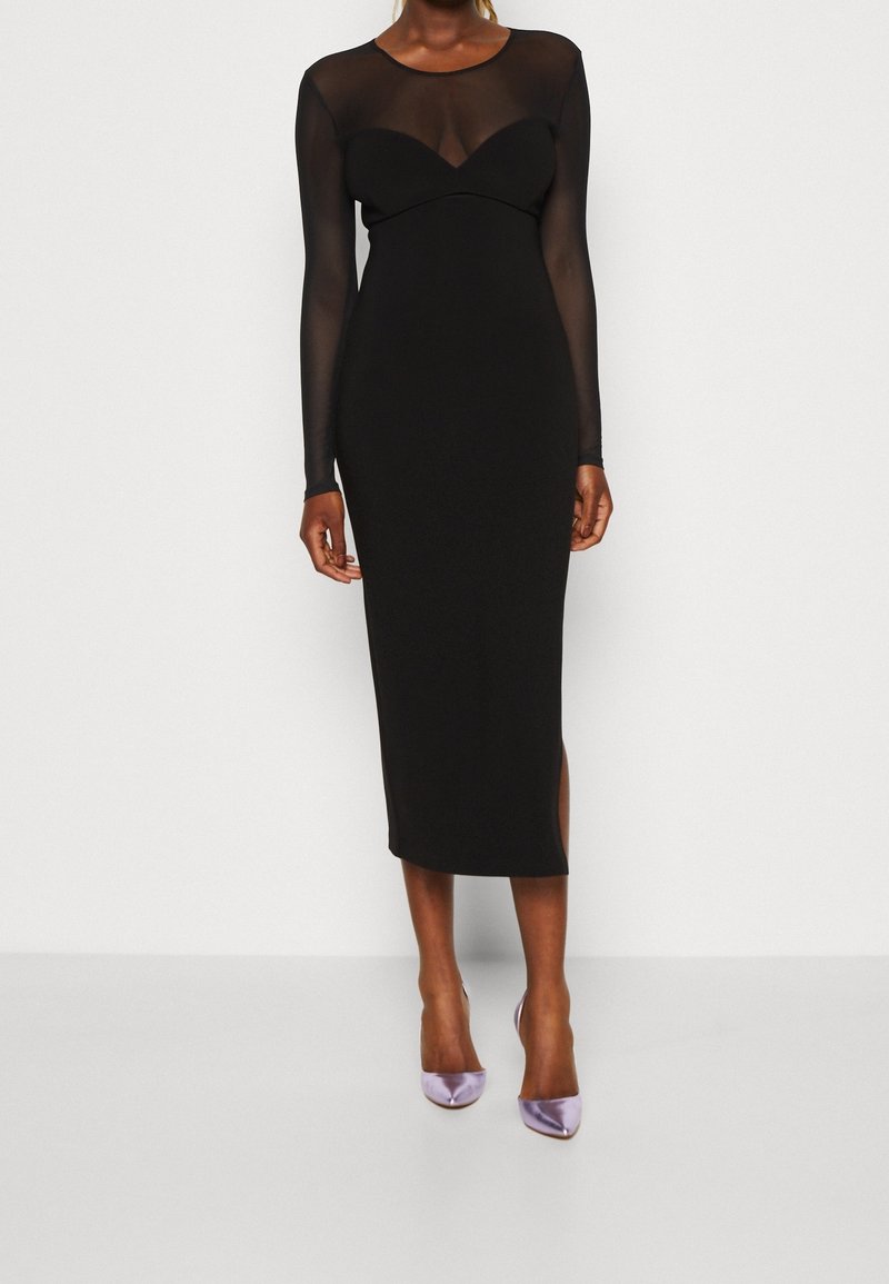 Czarna midi sukienka z długimi przezroczystymi rękawami, dopasowaną górą i delikatnym rozcięciem z boku, w połączeniu z fioletowymi satynowymi szpilkami.