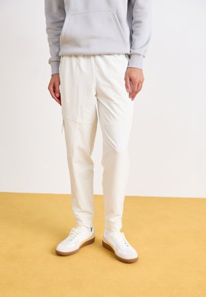 Pantalons de sport blancs avec une texture lisse, dotés d'une poche zippée et d'une coupe fuselée, associés à des baskets blanches sur un sol jaune.