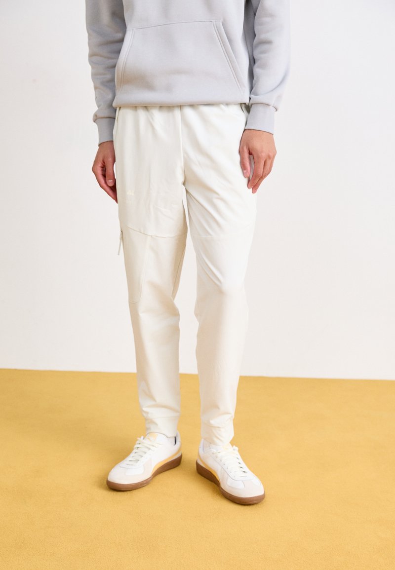 Witte sportbroek met een gladde textuur, voorzien van een ritszak en een tapse pasvorm, gecombineerd met witte sneakers op een gele vloer.