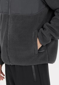 Harmaa fleece-takki, jossa on sileä yläosa, vetoketjullinen etuosa, sivutaskut ja teksturoitu pinta, yhdistettynä mustiin alaosiin.