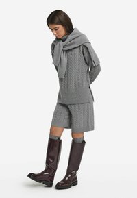 Grauer Kabelstrickpullover mit passenden Shorts, kombiniert mit einem grauen Schal. Das Model trägt hohe, bordeauxfarbene Lederstiefel mit einem eleganten Design.