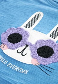 Hvid tegneseriekanin iført strukturerede lilla blomstermønstrede solbriller på blå stof med teksten "SMILE EVERYDAY".