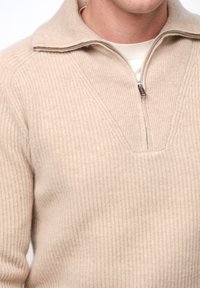 Beige ribbad stickad tröja med halvdragkedja och V-ringad design. Har en mjuk textur och en subtil glans från materialet.