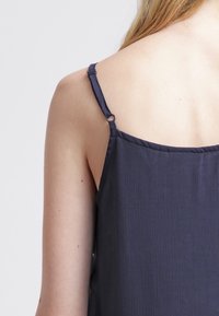 Granatowy bezrękawnik z teksturowanej tkaniny, z cienkimi ramiączkami. Ramiączko ma mały srebrny detal w postaci pierścionka na krawędzi.