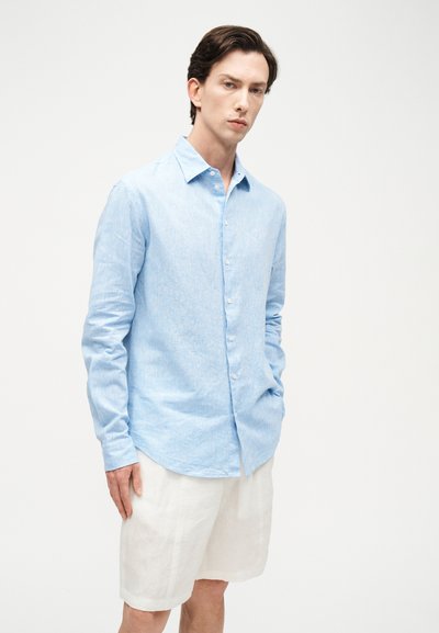 Νεαρός άνδρας που φορά ανοιχτό μπλε πουκάμισο με μακριά μανίκια και λευκά σορτς μέχρι το γόνατο, στέκεται με το ένα χέρι στην τσέπη.