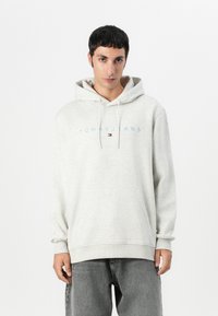Grå hettegenser laget av mykt stoff med en frontlomme og justerbar snøring. Har "TOMMY JEANS" i lyseblå tekst over brystet.