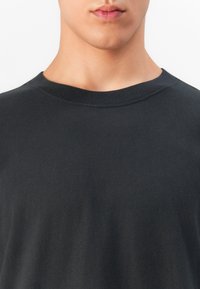 Adult tânăr îmbrăcat cu un tricou simplu de culoare închisă, care îi arată partea inferioară a feței, gâtul și partea superioară a pieptului, pe un fundal alb.