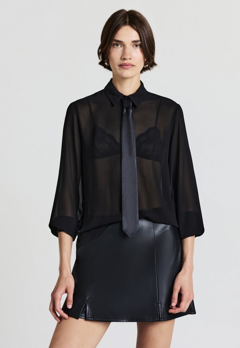 Blusa nera trasparente con colletto, legata con una cravatta nera elegante. Sotto, un reggiseno in pizzo nero visibile. Indossata con una mini gonna in pelle nera.