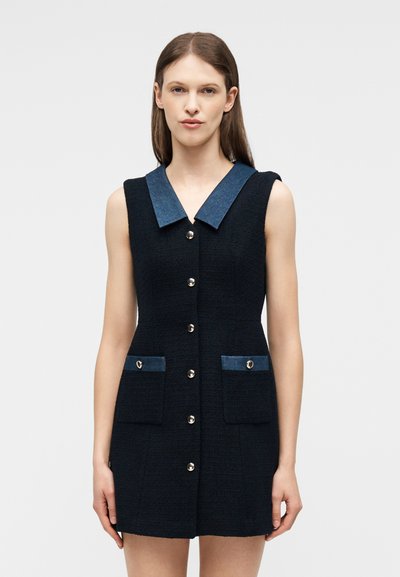 Γυναίκα που φορά αμάνικο μαύρο φόρεμα με σκούρο μπλε γιακά, κουμπιά μπροστά και δύο μπροστινές τσέπες, σε απλό ανοιχτό φόντο.