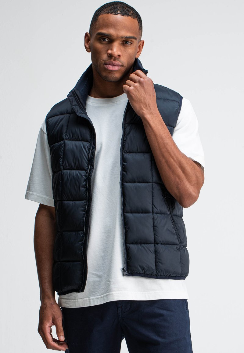 Schwarze Steppweste mit Reißverschluss vorne, gestepptem Design und hohem Kragen, getragen über einem hellgrauen T-Shirt mit lockerer Passform und Seitennähten.