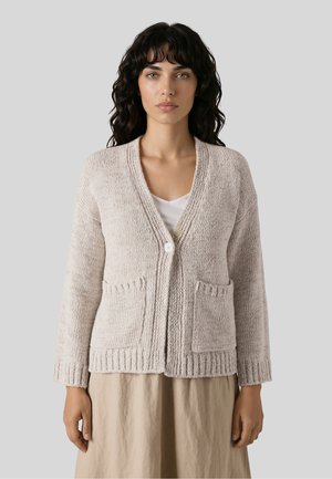 Kvinde med mørkt krøllet hår har en beige strikket cardigan med to forlommer og en enkelt knap over en hvid top og beige nederdel.