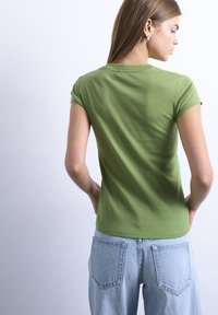 Γυναίκα με πράσινο κοντομάνικο πουκάμισο και ανοιχτό μπλε τζιν, κοιτάζοντας προς τα πίσω, με τα χέρια στη μέση, μπροστά σε απλό φόντο.