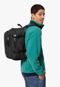 Musta reppu, jossa on useita vetoketjullisia lokeroita, säädettävät hihnat ja sivulukot, käytetty teal-värisen fleece-takin päällä.