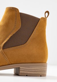 Stivaletto in pelle scamosciata giallo senape con pannello in tessuto marrone a coste, dettagli cuciti e tacco basso in legno per garantire stabilità.