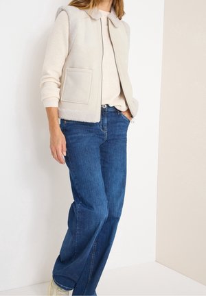 Frau trägt eine beige ärmellose Fleeceweste mit Reißverschluss über einem hellen Pullover, blaue Jeans und weiße Sneaker vor einem einfarbigen Hintergrund.