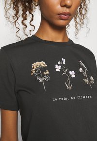 T-shirt noir avec des illustrations florales en beige, blanc et violet, avec le texte "pas de pluie, pas de fleurs" imprimé sous le design.