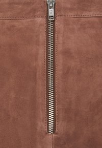 Tissu en suédine marron avec un détail de fermeture éclair métallique central. La fermeture éclair possède des dents en argent et une languette de marque, le tout sur une surface lisse.