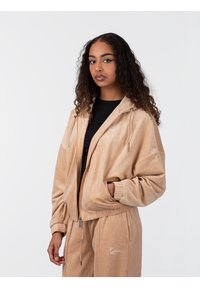 Chaqueta de ante beige con cremallera y capucha, puños acanalados y bolsillos frontales. Muestra una textura suave y un sutil logotipo en el pecho.