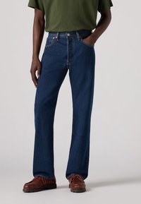Homme portant un jean droit bleu foncé, un t-shirt vert olive et des chaussures en cuir marron, debout avec une main dans la poche.