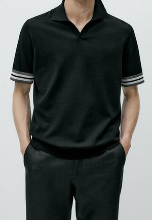 Mężczyzna w czarnym, krótkim polu dzianinowym z szarymi paskami na rękawach, z rękami w kieszeniach, na jednolitym jasnym tle.